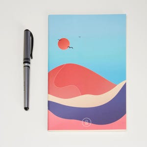 May include: A blue and pink notebook with a stylized illustration of a sunset over a desert landscape. The notebook has a small circle with the letters "O M" in the bottom right corner.