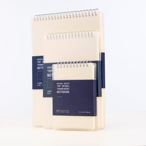 May include: Three spiral bound notebooks with clear plastic covers. The notebooks are beige and have a blue label with the text "Minimal Series Top Spiral Transparent Notebook".