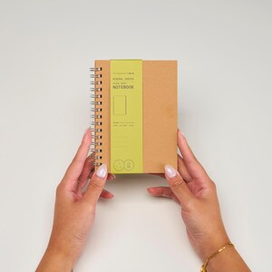 May include: A brown notebook with a green label that says "Designed by... Normal Notebook Spiral 96 Sheet Notebook".