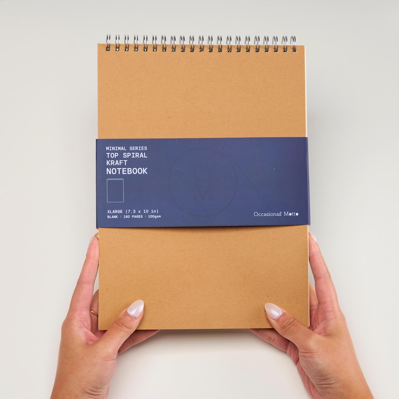 Op de afbeelding: Een bruin kraft notitieboek met een blauwe omslag waarop "Minimal Series Top Spiral Kraft Notebook" en "XLARGE (7.3 x 10 in) BLANK - 100 PAGES - 100gsm" en "Occasional Matte" staat.