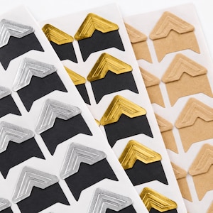 May include: Three vertical sheets of adhesive photo corners in silver, gold, and kraft paper brown. Each sheet features multiple V-shaped corner mounts arranged in rows, designed for securing photographs or documents onto a flat surface.