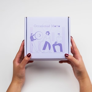 May include: A purple box with the text "Occasional Motto" on the front. The box features a graphic of three people, two women and one man, in a stylised illustration.