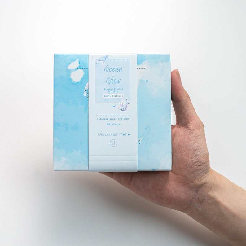 May include: A blue box with a white label that says "Ocean View" and features a design of clouds and seagulls. The label also says "Journal Stickers Gift Box" and "50 sheets".