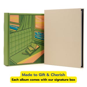 May include: A beige hardcover album standing next to a green rectangular box. The box features a graphic illustration of a train interior with a seat, a window view of a sunset, and an open book. The text Made to Gift & Cherish is visible.