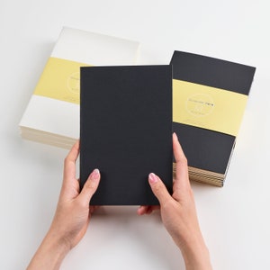 May include: Two stacks of notebooks, one with a black cover and the other with a white cover. Both covers have a yellow label with the text "Original Note" and a small blue "M" logo. A hand is holding a black notebook in front of the stacks.