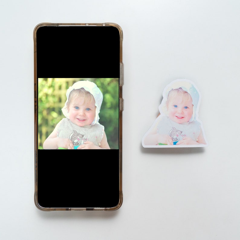 May include: A photo of a young child wearing a white bonnet and a light blue and white patterned shirt. The child is sitting in a grassy area and looking at the camera.