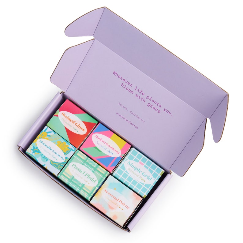 May include: A purple box with six small boxes inside. The small boxes are decorated with different patterns and colors, including green, blue, pink, and yellow. The text on the boxes reads: "Minimalist Greenery", "Stained Glass", "Modern Geometry", "Pastel Plaid", "Simple Grid", and "Seasonal Palette". The text on the larger box reads: "Wherever life plants you, bloom with grace. Irvine, California occasionalmotta"