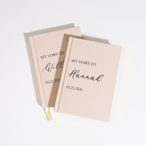 Op de afbeelding: Twee blushkleurige trouwboeken met de tekst "MY VOWS TO" gevolgd door een naam en de datum 03.23.2026 in zwart script. Elk boek heeft een crèmekleurig lint als bladwijzer.