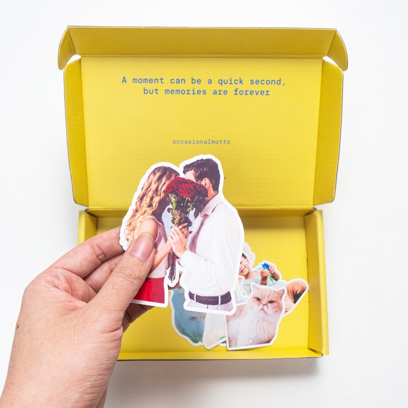 May include: A yellow box with a quote inside: "A moment can be a quick second, but memories are forever."  The box is open and a hand is holding a sticker of a couple kissing. The sticker is surrounded by other stickers, including a cat and a man holding a blue object.