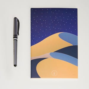 May include: A blue notebook with a stylized illustration of sand dunes under a starry night sky. The notebook has a small circle with the letters "OM" in the bottom right corner.