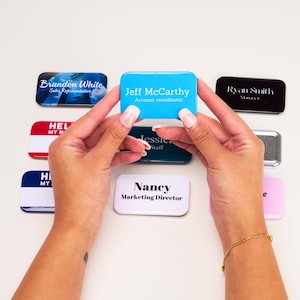 May include: A collection of colorful rectangular name tags with white text. The name tags are held in the hands of a person. The name tags read "Brandon White Sales Representative", "Jeff McCarthy Account Coordinator", "Ryan Smith Manager", "Hel My N", "Jessica Staff", "H My", "Nancy Marketing Director", and "e".