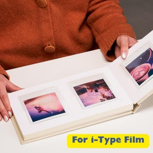 May include: A white photo album with a cream-colored interior and clear plastic sleeves for holding instant photos. The album is open to reveal three photos, one of which is a landscape shot of a lake and mountains, another is a photo of a person standing in a field, and the third is a photo of a cup of coffee. The album has the text "For i-Type Film" printed on the front.