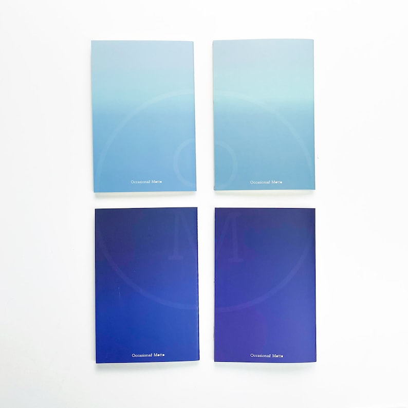 May include: Four small, blue and light blue notebooks with a gradient color design. Each notebook has the text "Occasional Marie" printed on the front.