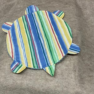 May include: A turtle-shaped fabric item with a striped pattern in blue, yellow, green, and red. The item has a circular body and four flipper-like appendages. The edges are finished with a decorative stitch, and the item is placed on a neutral-colored surface.