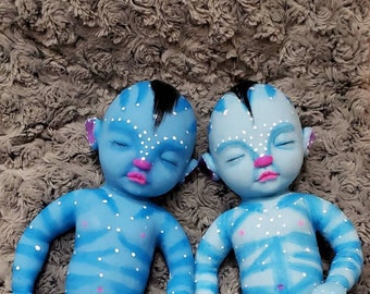 May include: Two blue silicone baby dolls with white dots and stripes, closed eyes, and pink lips. The dolls have black hair and are lying on a gray fuzzy surface.