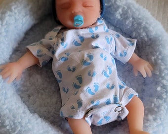 May include: A sleeping baby doll wearing a blue hat and a white onesie with blue footprints. The doll is lying in a blue fuzzy bed.