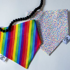 May include: Two dog bandanas. One features a rainbow striped pattern, the other has a white background with colorful polka dots. Both have a small white tag that reads "Sew Doggy Boutique". A black beaded necklace is draped across the bandanas.