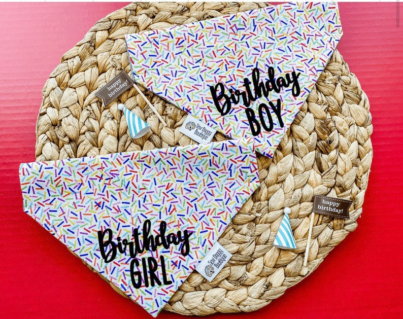 May include: Two triangular pet bandanas featuring a white background with a colorful sprinkle pattern. One bandana has the black text Birthday BOY, the other has Birthday GIRL. Each includes a small party hat and a brown flag labeled happy birthday!