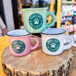 May include: Three small ceramic mugs in pastel colors: mint green, pink, and white. Each mug has a black rim and a curved handle. The mugs feature the text "PAWS OFF MY PUP CUP" around a paw print design. They are displayed on a wooden surface.