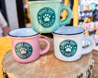May include: Three small ceramic mugs in pastel shades: mint green, pink, and white. Each mug has a black rim and a curved handle. The mugs feature the text "PAWS OFF MY PUP CUP" around a paw print design. They are displayed on a wooden surface.