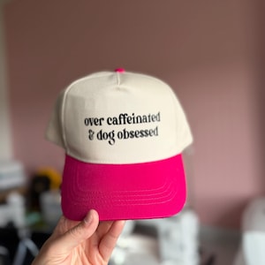 Op de afbeelding: Een baseballpet met een crèmekleurige kroon en een felroze klep. Op de voorkant van de kroon staat de zwarte tekst 'over caffeinated & dog obsessed' in een schreeflettertype. De pet heeft een klein roze knoopje aan de bovenkant in het midden.