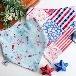 May include: A collection of patriotic-themed dog bandanas in red, white, and blue. One bandana features a light blue background with fireworks and stars. Two glittery star-shaped ornaments are also present.