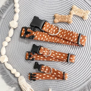 May include: Three brown and white dog collars with black buckles and silver D-rings. The collars are on a gray woven mat with a fringe border.