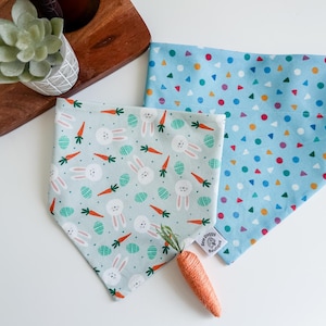 May include: Two triangular pet bandanas. One is light green with a pattern of white rabbits, orange carrots, and teal patterned eggs. The other is light blue with a pattern of multicoloured circles and triangles. A small fabric tag reads Sew Doggy Boutique.