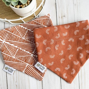 May include: Two dog bandanas with a rust orange background. One bandana has a geometric pattern in white, the other has a white repeating pattern of half circles. Both bandanas have a white label with the text "Sew Doggy Boutique".