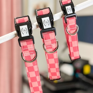 Pu&ograve; includere: Tre collari per cani a quadri rosa e bianchi con fibbie nere e anelli argentati. Ogni collare ha un'etichetta rettangolare nera con il testo "Sew Doggy Boutique" e una piccola silhouette di cane.