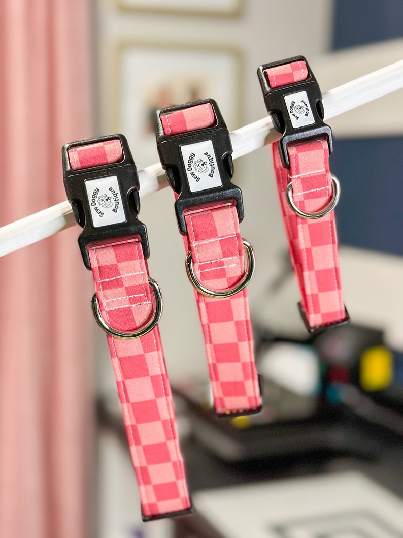 Pu&ograve; includere: Tre collari per cani a quadri rosa e bianchi con fibbie nere e anelli argentati. Ogni collare ha un'etichetta rettangolare nera con il testo "Sew Doggy Boutique" e una piccola silhouette di cane.
