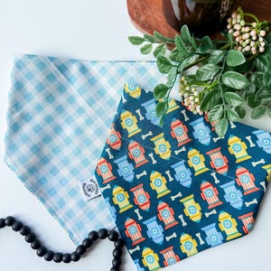 May include: Two dog bandanas. One is light blue gingham, the other is navy blue with a pattern of colorful fire hydrants and bones. A black beaded necklace is in the foreground. A wooden cutting board and a candle are in the background.