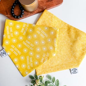 May include: Two yellow dog bandanas. One bandana has a repeating pattern of white suns and the text "SUNS OUT TONGUES OUT". The other bandana has a yellow swirl pattern. Both bandanas have a white tag that says "Sew Doggy Boutique".