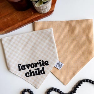 May include: Two dog bandanas. One is tan and white gingham with the words "favorite child" in black. The other is tan with white polka dots. Both are folded and placed on a white surface. A black beaded necklace is in the foreground.