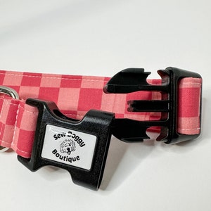 Pu&ograve; includere: Collare per cani a quadri rosa e bianco con una fibbia in plastica nera. La fibbia ha un'etichetta bianca con il testo "Sew Doggy Boutique" e un cane dei cartoni animati.