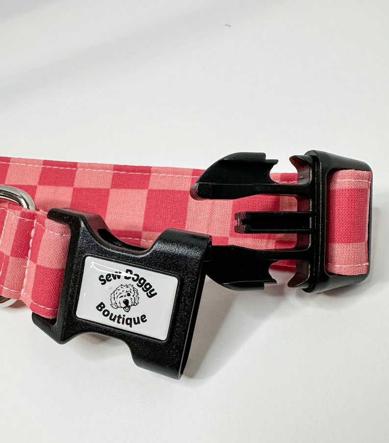 Pu&ograve; includere: Collare per cani a quadri rosa e bianco con una fibbia in plastica nera. La fibbia ha un'etichetta bianca con il testo "Sew Doggy Boutique" e un cane dei cartoni animati.