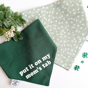 May include: Two dog bandanas. One is dark green with the text "put it on my mom's tab" in white. The other is light green with white polka dots. The bandanas are on a white surface with green shamrock decorations.