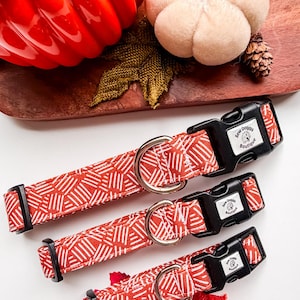 May include: Three dog collars in varying sizes featuring a red fabric with a white geometric pattern of short parallel lines. Each collar includes a black plastic side-release buckle with a Sew Doggy Boutique label and a silver metal D-ring.