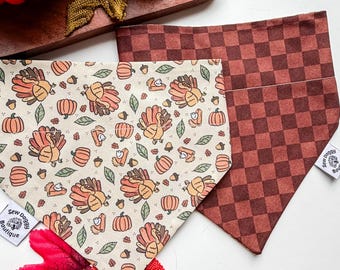 Op de afbeelding: Twee driehoekige bandana's voor huisdieren. De ene heeft een crèmekleurige achtergrond met een herhalend patroon van kalkoenen, pompoenen, eikels en taartpunten. De andere heeft een donkerbruin en roestkleurig ruitpatroon. Beide hebben een Sew Doggy Boutique label.