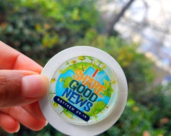 Puede incluir: Un soporte circular blanco para teléfono con un gráfico colorido. El gráfico presenta un diseño de globo terráqueo con las palabras "SHARE GOOD NEWS" y "MATTHEW 24:14". El fondo es una vista borrosa de follaje verde.
