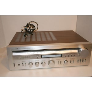 May include: An Akai AA-R50 stereo receiver with a silver metal front panel, featuring a horizontal radio frequency display, a signal strength meter, multiple control knobs, push buttons, a headphone jack, and a dark wood-grain top casing.