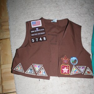 Puede incluir: Chaleco marr&oacute;n de Girl Scout con varios parches, incluyendo una bandera de Estados Unidos, Girl Scouts USA, Metro Detroit y el n&uacute;mero 5748. El chaleco tambi&eacute;n tiene varios parches triangulares con dise&ntilde;os coloridos.