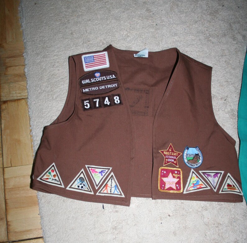 Puede incluir: Chaleco marr&oacute;n de Girl Scout con varios parches, incluyendo una bandera de Estados Unidos, Girl Scouts USA, Metro Detroit y el n&uacute;mero 5748. El chaleco tambi&eacute;n tiene varios parches triangulares con dise&ntilde;os coloridos.