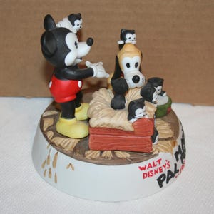 May include: A ceramic figurine of Mickey Mouse and Pluto with a group of kittens. Mickey is wearing red shorts and yellow shoes. Pluto is wearing a red collar. The kittens are black and white. The figurine is on a base with the text "Walt Disney's Pal" and a Mickey Mouse head.
