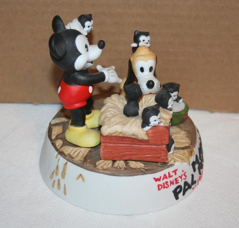 May include: A ceramic figurine of Mickey Mouse and Pluto with a group of kittens. Mickey is wearing red shorts and yellow shoes. Pluto is wearing a red collar. The kittens are black and white. The figurine is on a base with the text "Walt Disney's Pal" and a Mickey Mouse head.