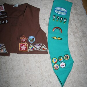 Puede incluir: Un chaleco marr&oacute;n de Girl Scout con varios parches y una faja de Girl Scout de color turquesa con parches y el texto "GIRL SCOUTS USA SOUTHEASTERN MICHIGAN" y el n&uacute;mero "5748".