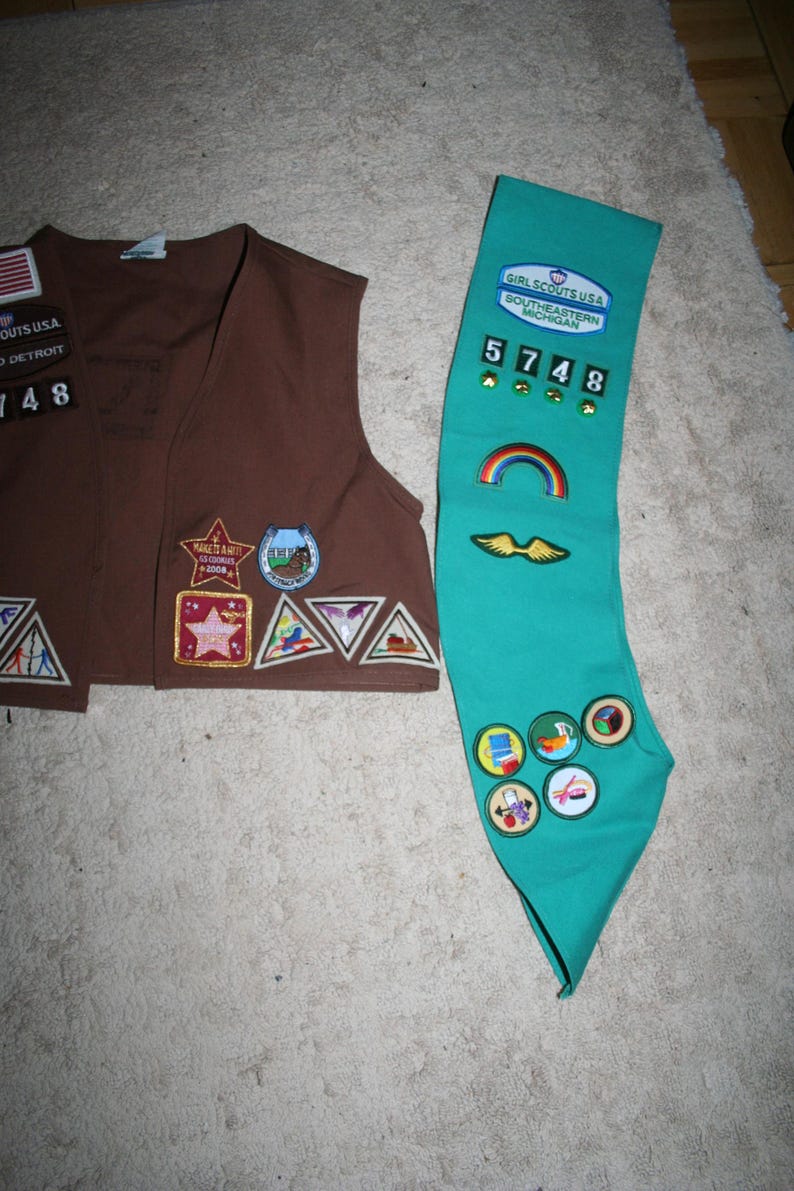Puede incluir: Un chaleco marr&oacute;n de Girl Scout con varios parches y una faja de Girl Scout de color turquesa con parches y el texto "GIRL SCOUTS USA SOUTHEASTERN MICHIGAN" y el n&uacute;mero "5748".