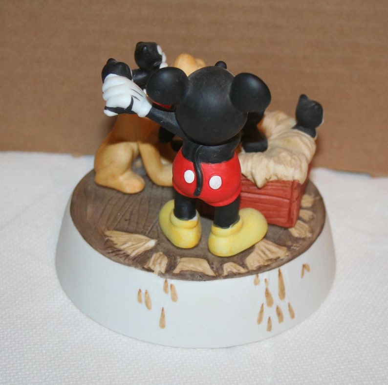 May include: A ceramic figurine of Mickey Mouse, Pluto, and Goofy. Mickey is wearing his signature red shorts and yellow shoes. Pluto is a brown dog with floppy ears. Goofy is a tall, yellow dog with a long snout. The figurine is set on a brown base with a white rim.