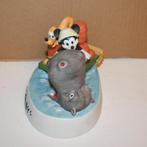 Puede incluir: Una figurita de cer&aacute;mica de Mickey Mouse, Pluto y un hipop&oacute;tamo. Mickey lleva un sombrero y sostiene una ca&ntilde;a de pescar. Pluto est&aacute; en la parte trasera del bote. El hipop&oacute;tamo est&aacute; en el agua. La figurita est&aacute; sobre una base que dice "Hip Hip Hooray!"
