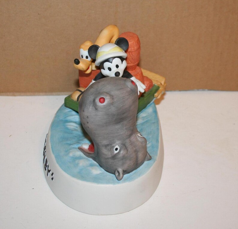 Puede incluir: Una figurita de cer&aacute;mica de Mickey Mouse, Pluto y un hipop&oacute;tamo. Mickey lleva un sombrero y sostiene una ca&ntilde;a de pescar. Pluto est&aacute; en la parte trasera del bote. El hipop&oacute;tamo est&aacute; en el agua. La figurita est&aacute; sobre una base que dice "Hip Hip Hooray!"